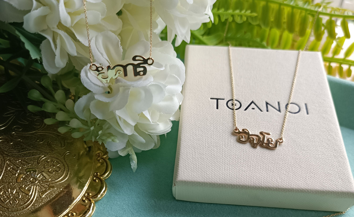 Two gold Thai name necklaces beside a Toanoi gift box